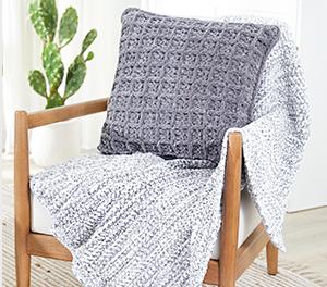 Image of a wooden chair that has a light gray knit blanket and gray pillow that is propped up on the back of the chair.