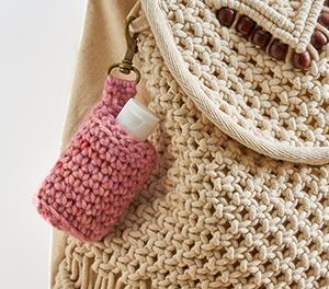 Image of a light pink crochet hand sanitizer key chain affixed to a tan crochet purse.