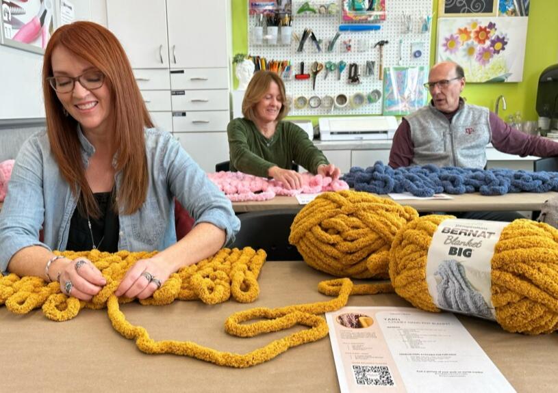 Three people make hand-knitted blankets with jumbo yarn in a Michaels classroom.