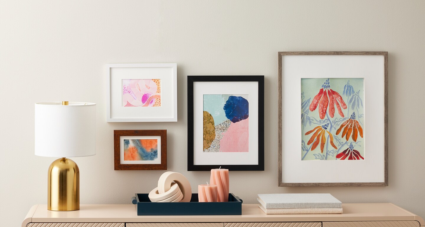 Contemporary frames arranged with a gold lamp, table decor, and books on a console table in front of a neutral background.