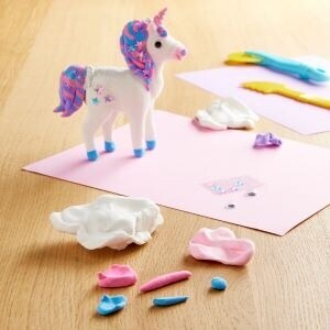 Small unicorn toy standing next to pieces of clay on pink construction paper. 