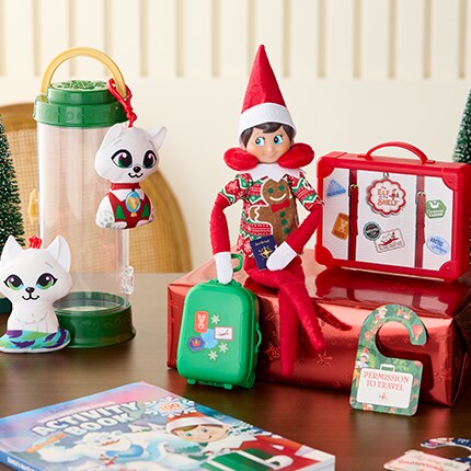 Elf sitting on a present with luggage in hand and in a neck pillow. (The Elf on the Shelf® Scout Elf® Carrier Limited 20th Anniversary Edition Kit)