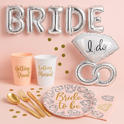 Bridal party decor set on pink background with "BRIDE" balloon letters, an "I do" ring balloon, cups labeled "Getting Drunk" and "Getting Married," plates with "Bride to Be," gold cutlery, and confetti.