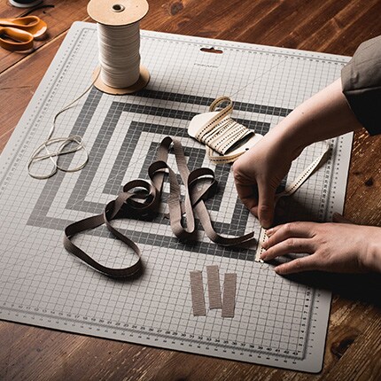 Hands weave ribbon on a gridded cutting mat. Nearby are a spool of string, measuring tape, scissors, and cut ribbon, evoking a meticulous crafting scene.