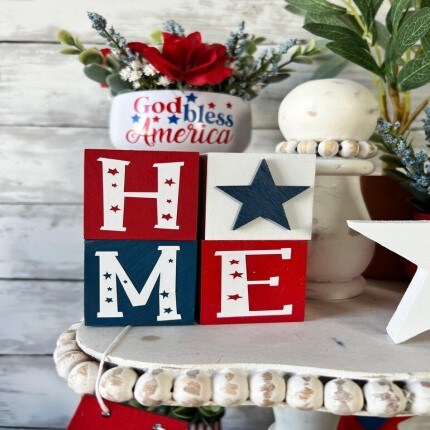 MakerPlace by Michaels Patriotic items on sale. Decorative blocks spell "HOME" in red, white, and blue with stars, evoking a patriotic theme. A vase with red flowers and "God bless America" text is in the background.