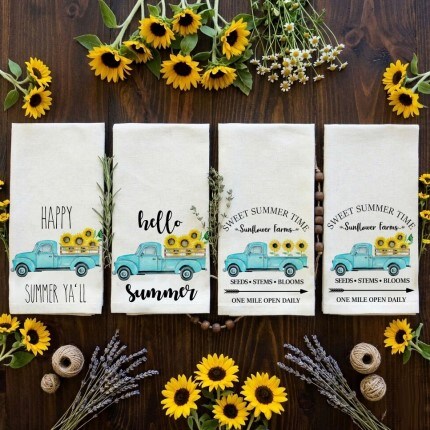 MakerPlace by Michaels Four decorative towels with blue vintage trucks and sunflowers lay on a wooden surface, framed by sunflowers and lavender sprigs, exuding a summer vibe.