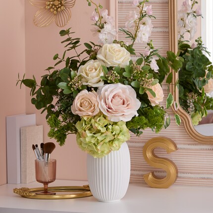  Floral arrangement in a white ribbed vase featuring cream and blush roses, green hydrangeas, and assorted greenery on a white surface.