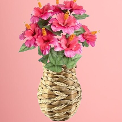 Woven basket vase holding a bouquet of vibrant pink hibiscus flowers with green leaves against a solid pink background.