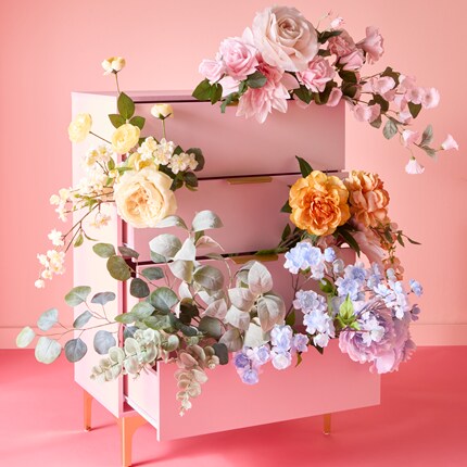 Pink chest of drawers with multiple drawers open, each filled with various stems including roses, peonies, and eucalyptus with a soft pink background.
