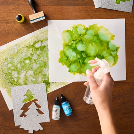 hand spraying green alcohol into onto paper with tree cut outs