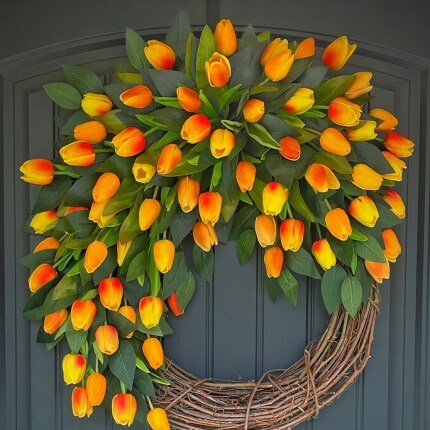 MakerPlace by Michaels A vibrant wreath of orange and yellow tulips with lush green leaves adorns a dark door. The tulips cascade from a rustic grapevine base, creating a cheerful and welcoming atmosphere.
