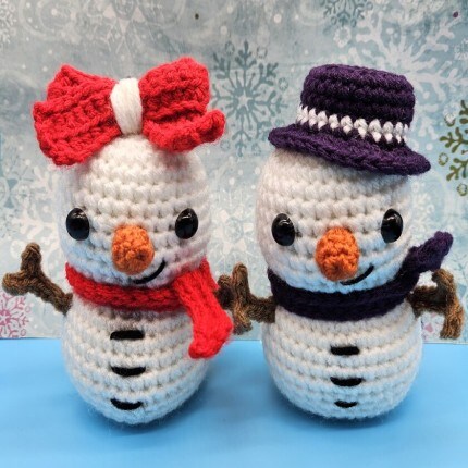 MakerPlace by Michaels Crafting & Fiber Arts. Two cheerful crocheted snowmen stand side by side on a blue surface, wearing a red bow and purple hat, with scarves, twig arms, and snowy patterned backdrop.