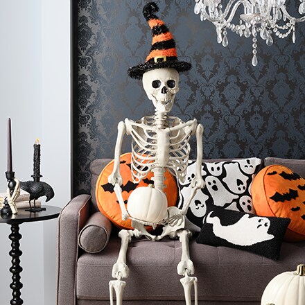 Decorative skeleton sitting in a couch with a black and orange striped hat and a white pumpkin in his hands.