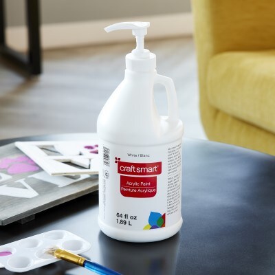 White pump bottle of 64 oz craft smart acrylic paint siting on black table next to paint pan and blue craft paint brush