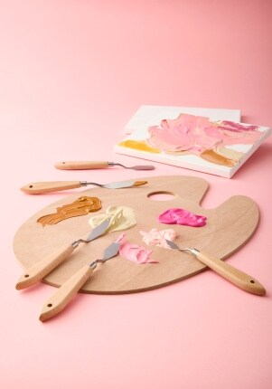 wood palette with paint and knives on pink background