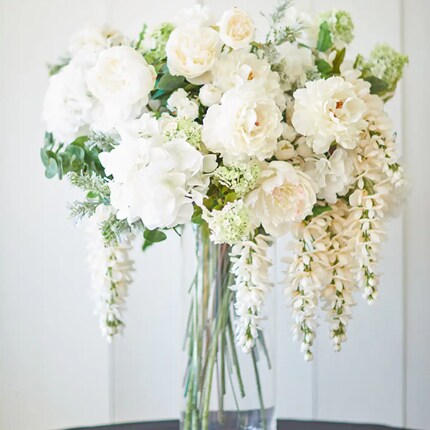 Floral arrangement featuring white roses, peonies, hydrangeas, and cascading wisteria in a clear glass vase. The bouquet showcases lush greenery and soft, delicate petals against a light, neutral background.