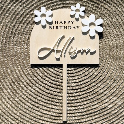 MakerPlace by Michaels Wooden cake topper with floral accents reads "Happy Birthday Allison." It lies on a textured, circular woven background, creating a rustic feel.
