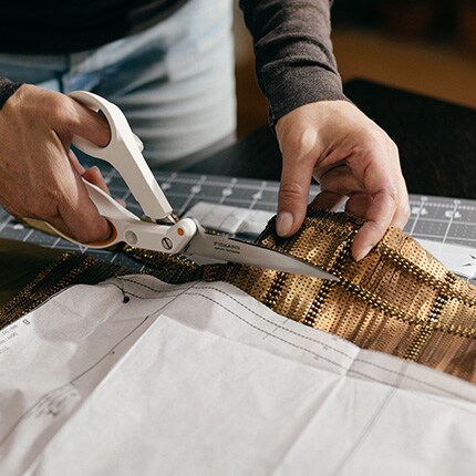 Hands cutting patterned fabric with scissors on a cutting mat, overlaid with a paper pattern. The scene conveys precision and craftsmanship.