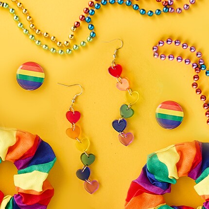 LGBTQ+ pride accessories arranged on a yellow background, including rainbow-striped scrunchies, heart-shaped dangling earrings in rainbow colors, rainbow bead necklaces, and round rainbow pins. 