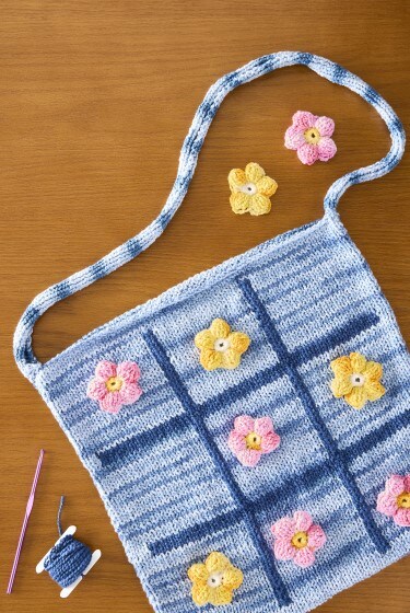 MakerPlace by Michaels Knitted blue and white tote bag with pink and yellow flower accents. Pink yarn, crochet hook, and spool of thread are nearby on a wooden table.