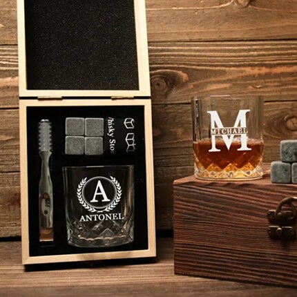 MakerPlace by Michaels A black gift set with a crystal glass, ice stones, and brush beside a whiskey tumbler on a wooden chest, evoking a refined, rustic mood.
