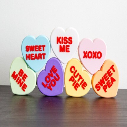 MakerPlace by Michaels Heart-shaped candies in pastel colors with red text: "SWEET HEART," "KISS ME," "XOXO," "BE MINE," "LOVE YOU," "CUTIE PIE," "SWEET PEA." Placed on a dark surface.