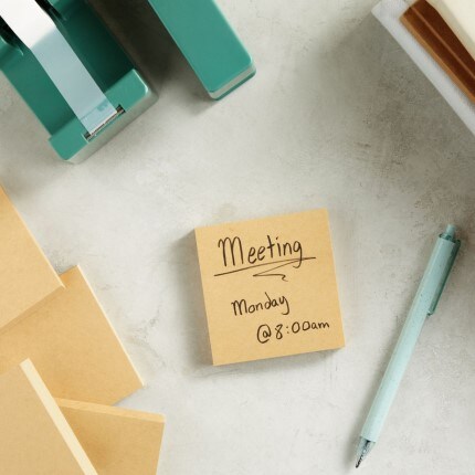 green stapler and tab dispenser sitting on table with sticky note that says "meeting Monday @ 8:00am"