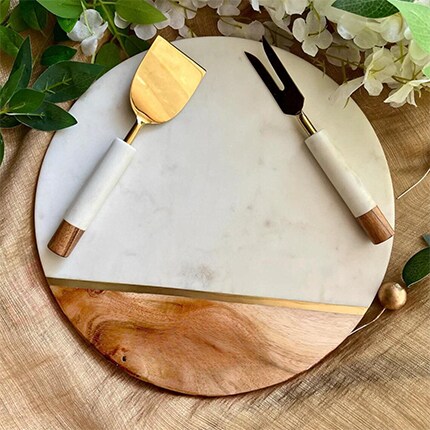 MakerPlace by Michaels A round marble-and-wood cheese board with gold serving knife and fork, framed by green leaves and white flowers in an elegant, inviting setting.