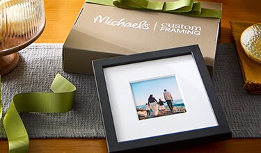 A black frame leaning on a Michaels custom framing box with a green bow on it.