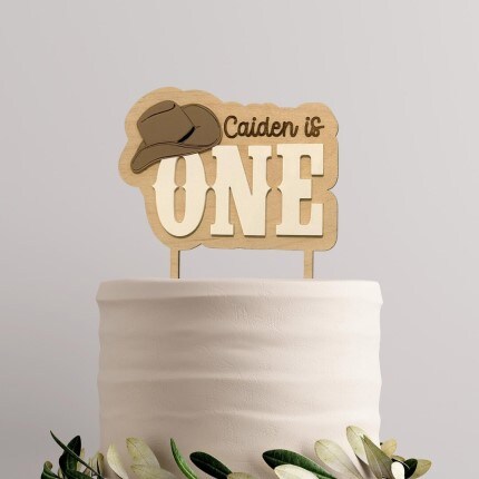 MakerPlace by Michaels Western-themed White cake topped with a wooden sign reading "Caiden is ONE," featuring a cowboy hat. Green and white leaves decorate the cake base. Festive tone.