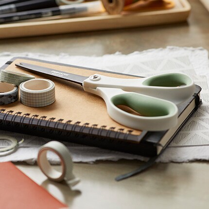 Scissors with green handles rest on a spiral notebook, surrounded by rolls of washi tape on a lace-covered table. The scene feels creative and organized.