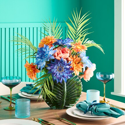 Vibrant floral centerpiece featuring blue, orange, and pink flowers with green palm leaves in a large green vase. 
