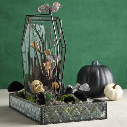 Decorative Halloween-themed terrarium with a glass and metal frame contains a small skull, black mushrooms, dried plants, and artificial butterflies on a mossy base. 