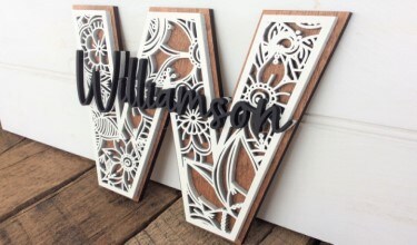 MakerPlace by Michaels Ornate wooden letter "W" with intricate white floral patterns, featuring the name "Williamson" in bold black script. Rustic and elegant decor.