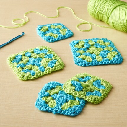 Crochet granny squares in blue and green are arranged on a wooden surface. A crochet hook and a ball of green yarn are nearby, creating a vibrant and crafty scene.