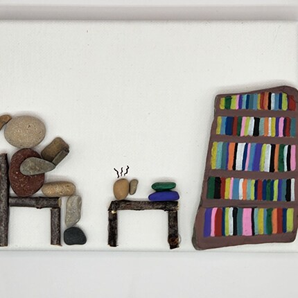 MakerPlace by Michaels Pebble art shows a cozy reading nook: a colorful bookshelf beside a tiny stone table with a steaming cup, chair, and stacked books, evoking calm warmth.