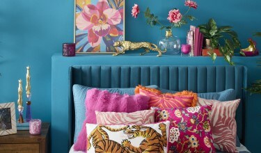 Pink Jungle Decor Collection: bedroom with blue walls, vibrant colors. Pillows in orange and pink colors over the bed. Potted Plants next to both sides of the bed. Tabletop decorations on the side table and over the bed frame.