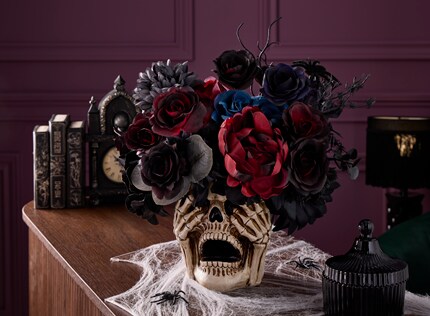 Decorative skull vase filled with dark red, black, and deep blue artificial flowers, placed on a wooden surface with white spiderwebs and plastic spiders. 