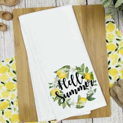 MakerPlace by Michaels White kitchen towel on a wooden board with "Hello Summer" and lemon design. Surrounded by lemon-patterned decor, evoking a fresh, cheerful vibe.