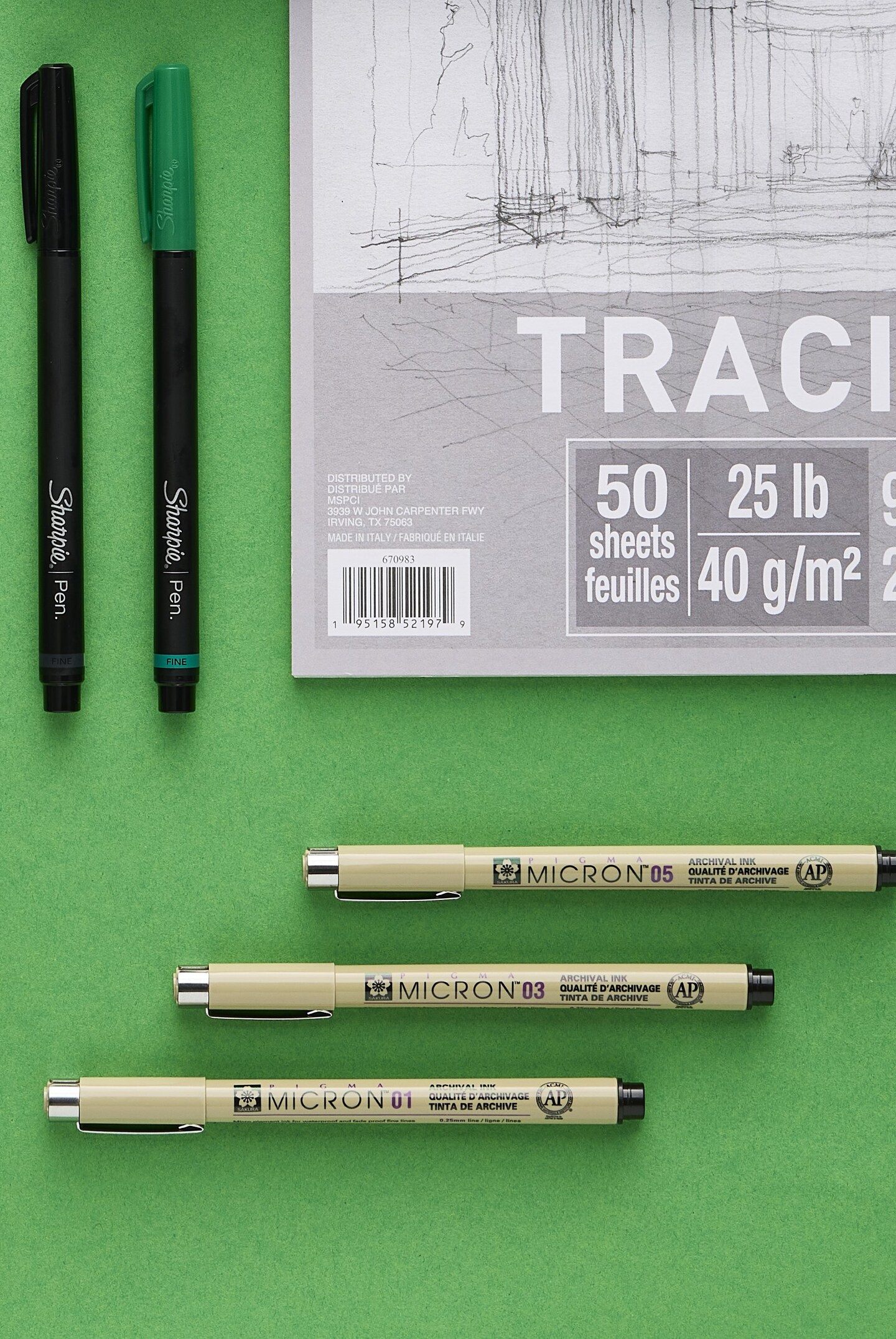Green & black sharpie pen laying next to each out. Pigma Micron pen sizes 01, 03, 05 pens laying side by side. Tracing paper displayed next to the pens over a green background. 