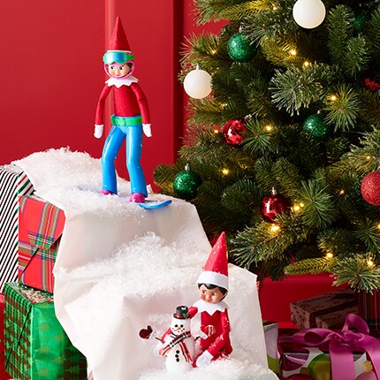 Elves sitting on a white felt sheet, "snowboarding" down a stack of presents. 