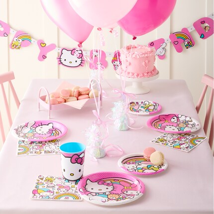 A colorful Hello Kitty-themed birthday setup. Features include a cake with pink icing, cupcakes with candles, character plates, napkins, cups, and balloons. Playful and festive atmosphere.