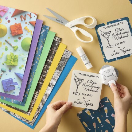 assorted scrapbook sheets laying on yellow background next to white scissors, glue stick, and paper punch with a hand holding an invitation 