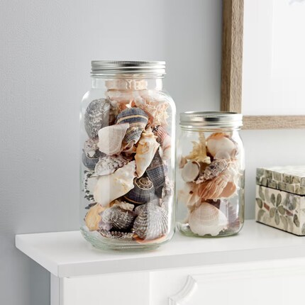 two clear glass jars filled with assorted seashells placed on a white mantelpiece.
