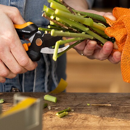Hands holding orange mesh bag and trimming green stems with black pruning shears on a wooden table. The scene conveys focus and care in gardening.