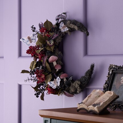 Moon-shaped wreath decorated with red flowers, green leaves, mushrooms, and white butterflies hanging on a lavender paneled wall.
