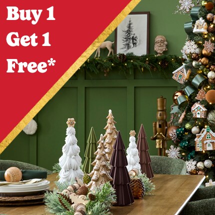 green room with reindeer and stockings on wall, with decorated Christmas tree and wood table with ceramic tree accents with Buy 1 Get 1 Free off in white text on red triangle