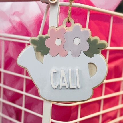 MakerPlace by Michaels A pastel teapot-shaped decoration with "CALI" text, adorned with pink and purple flowers, hangs on a white wire basket against a pink background.