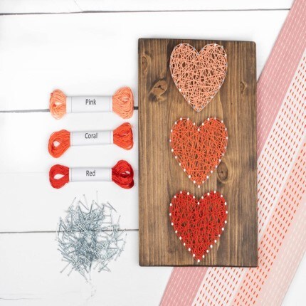MakerPlace by Michaels Three heart designs in pink, coral, and red string art on wood. Thread spools labeled by color are on the left, with nails below. A DIY craft setup.