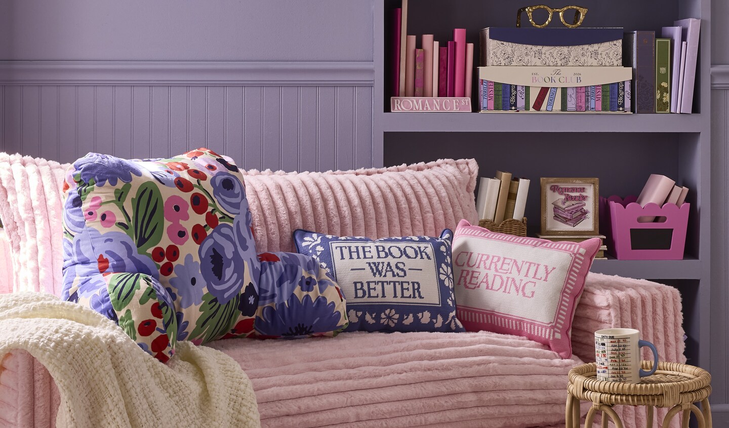Next Chapter Décor Collection: Pink couch with pink and purple pillows with book-related quotes. There's a purple shelf at the back that has pink and purple books, as well as some decorative boxes in the same colors.